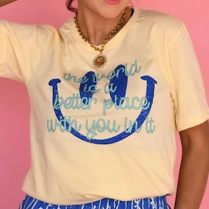 May include: A light yellow t-shirt with a blue smiley face and the text "the world is a better place with you in it." The person is wearing blue and white striped shorts.