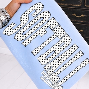 May include: A light blue sweatshirt with the word "SOFTBALL" in large, black polka-dotted letters. The text "hippydecorandmore" is below the main text. Additional text is visible on the left side of the sweatshirt.