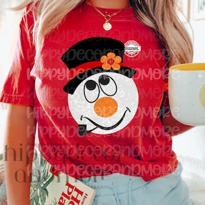 May include: Red t-shirt with a snowman graphic. The snowman is wearing a black top hat with a red band and a red flower. The snowman has a white face with black eyes, a carrot nose, and a black pipe.