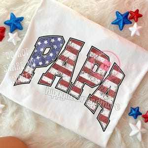 Puede incluir: Camiseta blanca con la palabra "PAPA" en letras de bandera estadounidense roja, blanca y azul desgastadas.