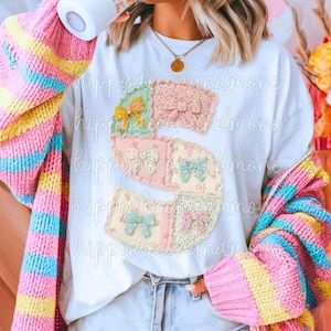 May include: White t-shirt with a patchwork letter "S" design, adorned with floral patterns and bows. The model wears a colourful striped cardigan in pink, yellow, and blue. A gold pendant necklace is also visible.