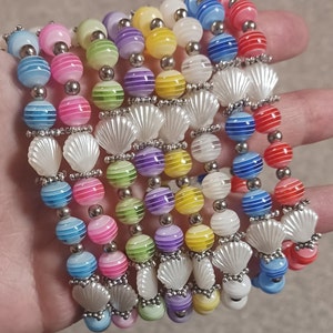 May include: A collection of colorful beaded bracelets. Each bracelet features a mix of striped, round beads in various pastel colors, interspersed with white seashell-shaped beads. The bracelets are held together with silver-toned metal accents.