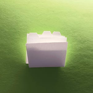May include: A stack of white file folders with tabs on the top edge. The folders are against a green background.