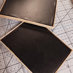 May include: Two rectangular, black, textured trays with light brown wooden frames. The trays are positioned on a cutting mat with a grid pattern. The black surfaces appear to be made of a durable material.