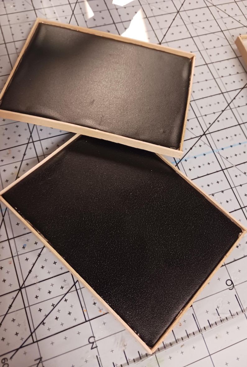 May include: Two rectangular, black, textured trays with light brown wooden frames. The trays are positioned on a cutting mat with a grid pattern. The black surfaces appear to be made of a durable material.