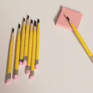 May include: A collection of yellow pencils with black tips and pink erasers. One pencil rests on a stack of pink sticky notes. The pencils are arranged on a white surface.