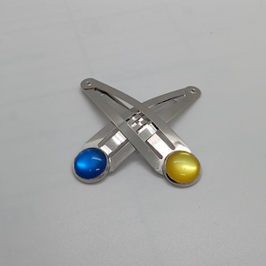 May include: Two silver hair clips with a blue and yellow gem on each. The clips are crossed over each other.