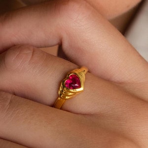 Puede incluir: Un anillo de oro con una piedra preciosa roja en forma de corazón. El anillo está en el dedo de una persona.