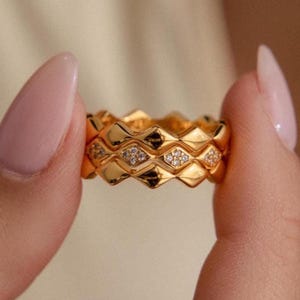 Puede incluir: Un anillo dorado con un patrón de diamantes repetido y pequeñas piedras transparentes. El anillo se sostiene entre dos dedos, mostrando su intrincado diseño y acabado pulido. Esta joya es un accesorio de moda.