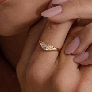 May include: A gold-colored ring with a curved design, set with small, clear gemstones. The ring is worn on a finger, showcasing its delicate details and elegant style. The ring's design is a subtle yet eye-catching piece of jewelry.