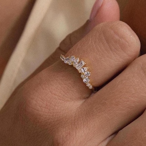 Puede incluir: Un anillo dorado con múltiples piedras preciosas transparentes, engastadas en un diseño curvo. El anillo está en el dedo de una persona, con la mano y el brazo visibles. El diseño del anillo presenta piedras redondas y rectangulares.