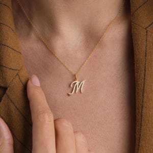 Puede incluir: Un collar de cadena de oro con un pequeño colgante de letra "M" cursiva incrustado con piedras de circonita cúbica transparentes.
