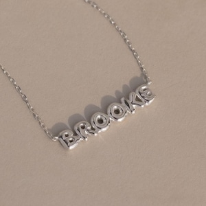 May include: A delicate silver necklace featuring the name "Brooke" in a block letter font. The necklace has a fine chain and is displayed on a neutral background, highlighting the personalized pendant.