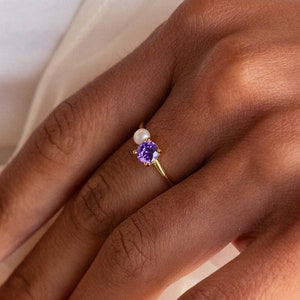Peut inclure: Une bague en or avec une pierre précieuse violette et une perle blanche. La bague est portée sur un doigt.