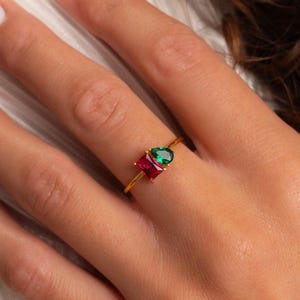 Puede incluir: Un anillo de oro con dos piedras preciosas distintas: una piedra de color rojo intenso con corte rectangular y una piedra de color verde vibrante con forma de pera, montadas una al lado de la otra en una banda de oro fino y pulido.