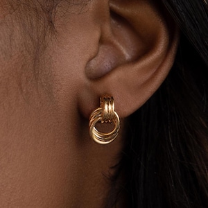 Puede incluir: Pendientes de nudo en tono dorado. Los pendientes son pequeños y delicados, con un diseño sencillo.