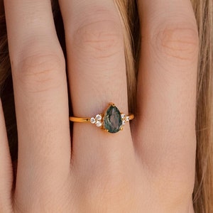 May include: A gold ring featuring a pear-shaped, dark green gemstone. The ring is accented with small, round, white gemstones on either side of the main stone. The band is a thin, polished gold.