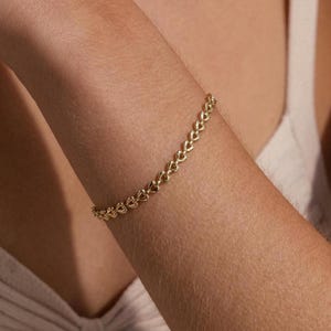 May include: A gold-colored bracelet with a delicate chain design is displayed on a person's arm. The bracelet is made of small, interconnected links. The arm is in front of a light-colored top.