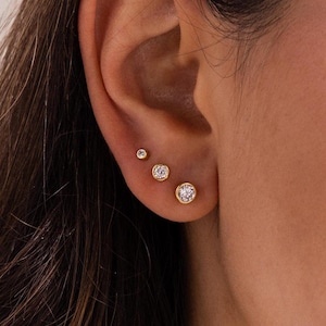 Puede incluir: Tres pendientes de oro con gemas redondas y transparentes. Los pendientes son de tamaños graduados, siendo el más grande el de abajo. Los pendientes están engastados en un engaste de bisel dorado. Los pendientes se muestran en una oreja.