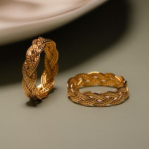 May include: Two gold-coloured braided rings with a detailed woven design are on a light green surface. A white teacup and saucer are in the background, out of focus.