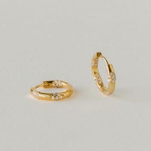 May include: A pair of small gold hoop earrings featuring a textured surface with embedded clear stones. One earring lies flat while the other stands upright, showcasing the circular design and the sparkling accents set into the metal band.