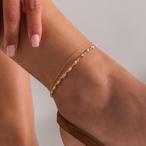 Puede incluir: Juego de joyas doradas. Una pulsera con pequeños adornos redondos adorna la muñeca, mientras que una delicada tobillera con detalles similares rodea el tobillo. Las joyas complementan el tono de la piel y la ropa de color neutro.