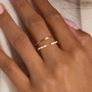 May include: A gold-colored open ring with small, rectangular, white gemstones. The ring is worn on a hand with manicured nails. The background is a soft, neutral color.