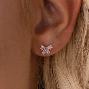 May include: A close-up of a small, bow-shaped stud earring. The earring is made of small, clear stones, likely cubic zirconia or diamonds, set in a silver or white gold metal. The earring is on a person's ear.