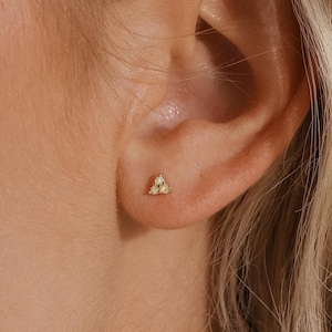 Könnte beinhalten: Nahaufnahme eines Ohrs mit einem kleinen Ohrstecker aus Gold. Der Ohrring hat ein dreieckiges Design mit drei kleinen, funkelnden Steinen. Die Person hat blondes Haar.