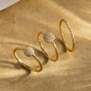 May include: Four gold rings with clear stones. Three of the rings have a square-shaped stone with smaller stones surrounding it. The fourth ring has a row of small stones.