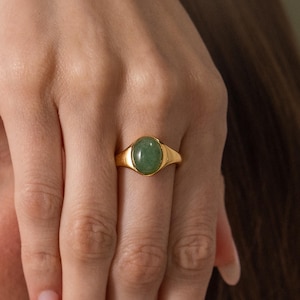 May include: A gold ring featuring a smooth, oval-shaped green stone set in a polished metal band. The ring is worn on the middle finger, showcasing the vibrant green gemstone and the warm, reflective quality of the gold-toned metal setting.