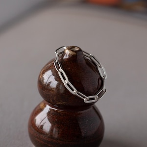 May include: A silver chain bracelet rests on a small, dark brown, gourd-shaped ceramic vase. The vase has a glossy finish and sits on a gray surface, with a blurred open book in the background.