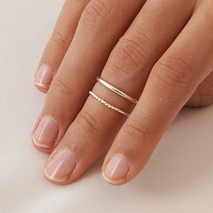 May include: Two silver rings on a finger. The top ring is a simple band, and the bottom ring is a delicate chain.