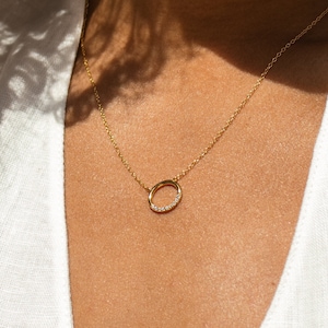 May include: A gold chain necklace with a small circle pendant encrusted with tiny diamonds. The necklace is worn by a person with brown skin and curly hair.