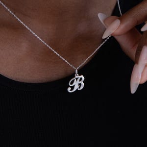 Puede incluir: Un collar plateado con un colgante con la letra "B" en cursiva. El collar está sobre una cadena fina. La persona lleva una camiseta negra.