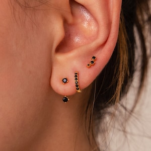 May include: A close-up of a person's ear with three gold earrings featuring black stones. The earrings are a small stud, a curved bar with stones, and a dangling earring with a small stone at the end.