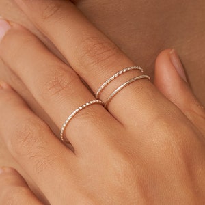 May include: A close-up of a hand wearing three thin silver rings. The rings are stacked on the ring finger. The rings are simple and delicate.