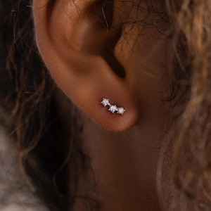May include: A close-up of a silver ear cuff with three small, sparkling gemstones. The ear cuff is curved to fit the ear's natural shape. The gemstones are clear and set close together, creating a subtle yet elegant look. The jewelry is worn on the ear.