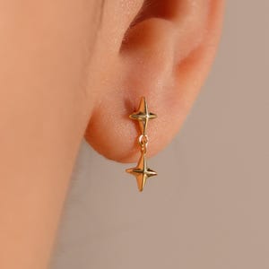 Puede incluir: Primer plano de pendientes colgantes dorados con forma de estrella. Cada pendiente tiene dos estrellas conectadas, una encima de la otra. Los pendientes se muestran en detalle, destacando su delicado diseño y el tono cálido del metal.