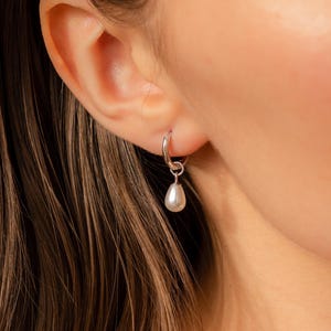 Puede incluir: Un pendiente de aro plateado con una sola perla blanca colgando.