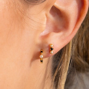 Könnte beinhalten: Ein Paar goldene Creolen mit schwarzen und weißen Steinen. Die Ohrringe sind klein und zart und wären ein tolles Geschenk für einen Freund oder eine geliebte Person.