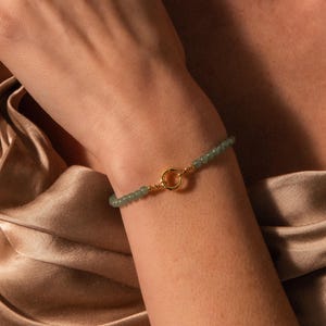 May include: A beaded bracelet featuring small, round, sage green beads strung on a cord, connected by a circular gold-toned metal clasp at the center. The bracelet is worn around the wrist, showcasing the contrast between the green beads and the gold.