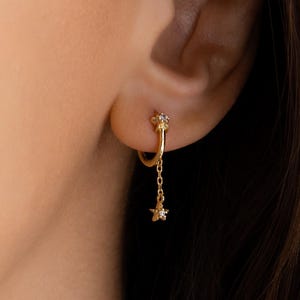 May include: A gold-coloured earring featuring a small hoop design with a dangling chain. The chain holds a small star-shaped charm. The earring is worn on the ear of a person with long, dark hair.