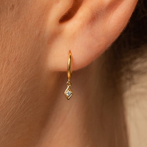 Close up on ear. A gold huggie hoop earring is in the the first lobe piercing. The hoop earring has a dangling charm in a dainty diamond shape with a circular diamond stone nestled inside it.