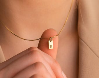 May include: A gold-toned necklace featuring a thin chain and a small, rectangular pendant charm. The pendant is engraved with the capital letter R. The necklace is worn around the neck, with the pendant resting near the collarbone area.