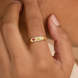 A gold signet ring with birthstones is on the pointer finger. There is a Ruby heart stone, a Topaz baguette stone, and a clear crystal circle stone next to each other.