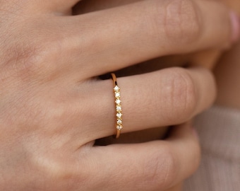 May include: A thin gold ring featuring a row of five small, evenly spaced, star-shaped accents along the top of the band. The ring has a polished finish and is worn on the ring finger, showcasing a delicate and slender design with a metallic lustre.