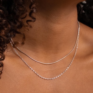 May include: A close-up of a person wearing two silver necklaces. The necklaces are layered, with a thin chain on top of a thicker chain. The person is wearing a brown off-the-shoulder sweater.