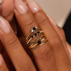 以下が含まれることがあります： 黒と白のダイヤモンドがついた、ゴールドのリングを5つ重ねてつけた手。リングは繊細で、シェブロン、スクエア、小さなダイヤモンドの集まりなど、さまざまなデザインが施されています。