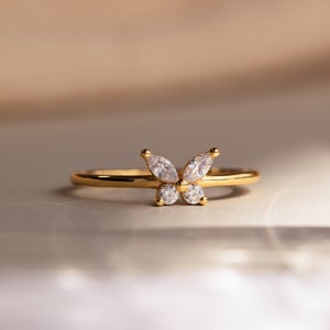 A close up of our Mariposa Butterfly Ring in 18K Gold finish placed on a white table and a clear glass vase in the foreground - featuring an about 5.5mm x 8mm CZ Diamond Butterfly Charm and an about 1.5mm Thin Ring Band.